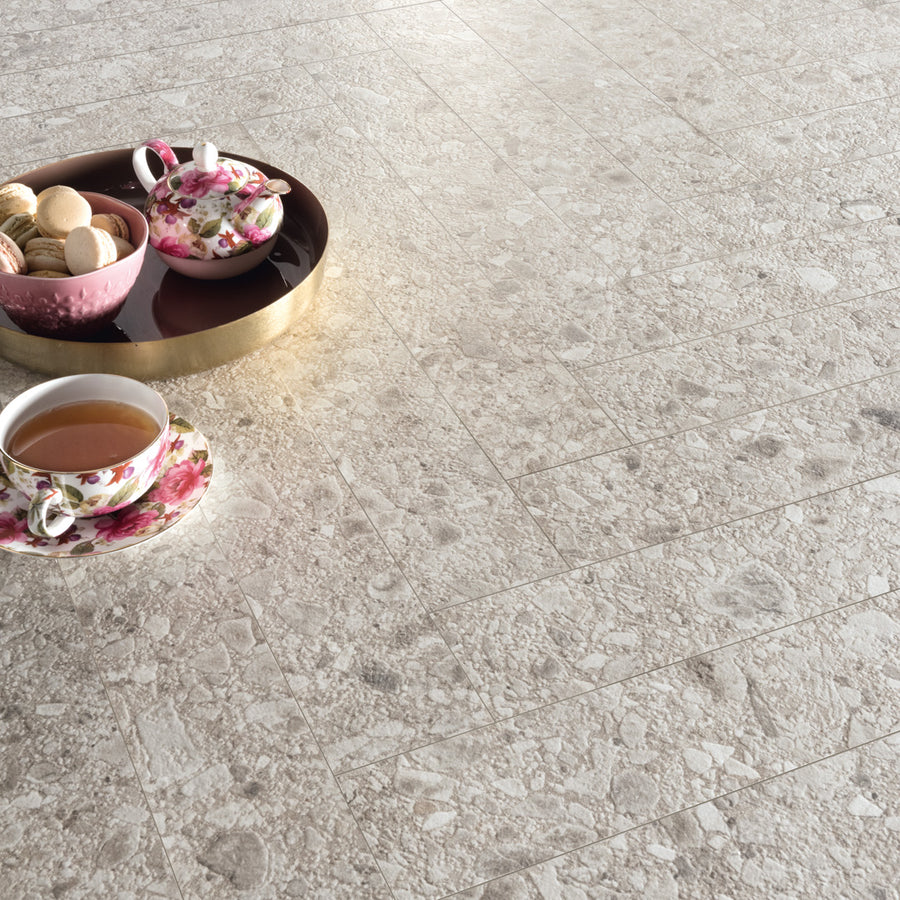 A floral teacup filled with tea, a teapot, and a bowl of cookies rest on a round tray on a textured stone tile floor.