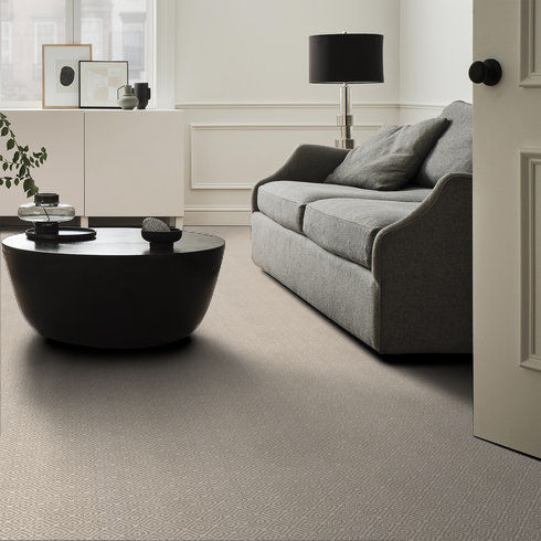 A modern living room with a gray sofa, a round black coffee table, a white sideboard with decor, a black table lamp, and neutral-colored walls and carpet. Natural light streams in from a window.