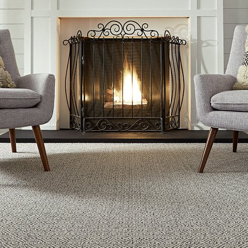 A cozy living room with two gray armchairs facing a lit fireplace. The fireplace has an ornate black screen, and the floor is covered with a light textured carpet.