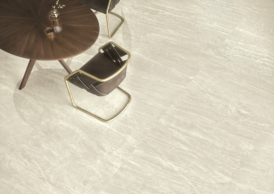 A round wooden table with two modern brown chairs sits on a floor of light gray, textured tiles. A glass vase with white flowers and a cup are on the table.