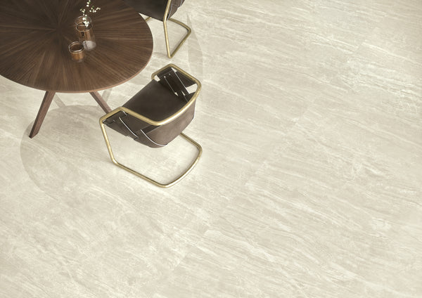A round wooden table with two modern brown chairs sits on a floor of light gray, textured tiles. A glass vase with white flowers and a cup are on the table.
