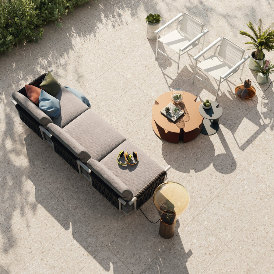 A modern outdoor patio set with a beige cushioned sofa, two white chairs, and round coffee tables on a tiled floor, surrounded by potted plants and decorated with throw pillows and a pair of sandals.