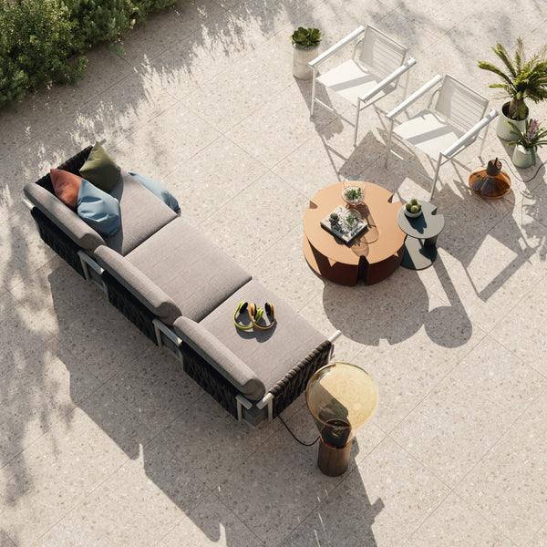A modern outdoor patio set with a beige cushioned sofa, two white chairs, and round coffee tables on a tiled floor, surrounded by potted plants and decorated with throw pillows and a pair of sandals.