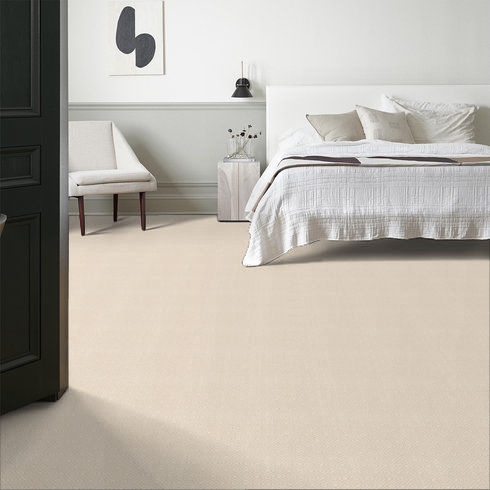 Minimalist bedroom with a white bed, neutral bedding, a small white chair, a nightstand with a vase and lamp, modern wall art, and beige carpet flooring. Walls are light with simple trim detailing.