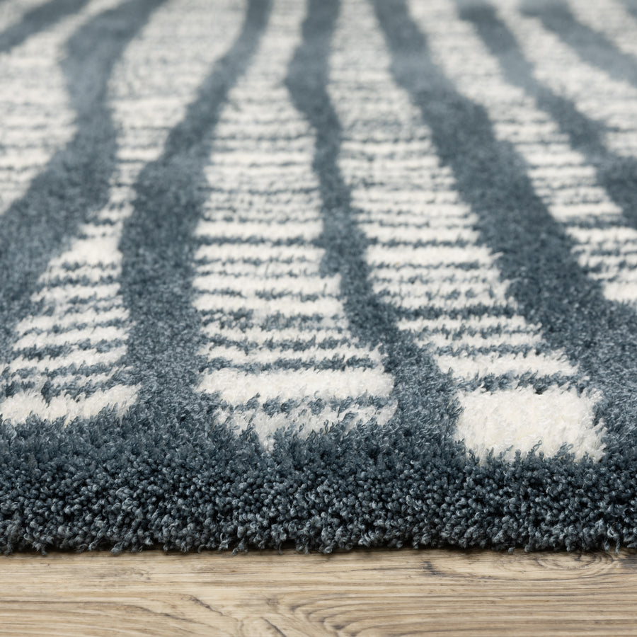 Close-up of a textured area rug with alternating blue and white striped patterns, placed on a wooden floor. The rug appears soft and plush.