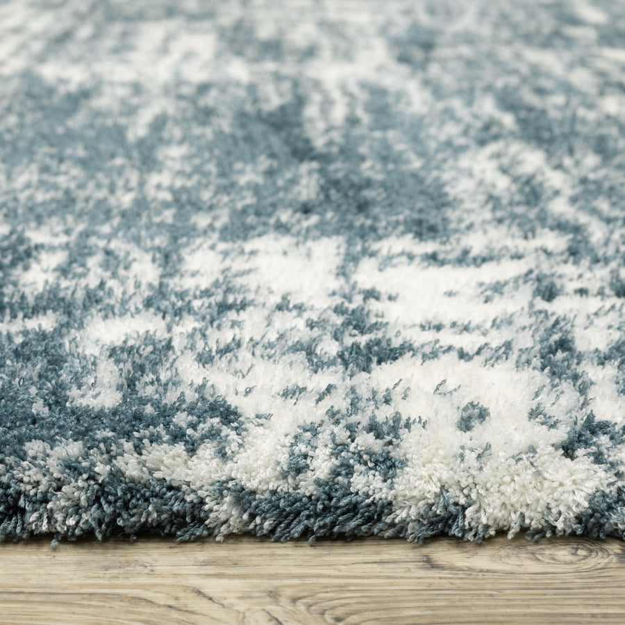 Close-up of a plush area rug with a blue and white abstract pattern, showing its soft, shaggy texture. The rug is placed on a wooden floor.