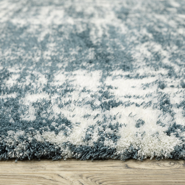 Close-up of a plush area rug with a blue and white abstract pattern, showing its soft, shaggy texture. The rug is placed on a wooden floor.