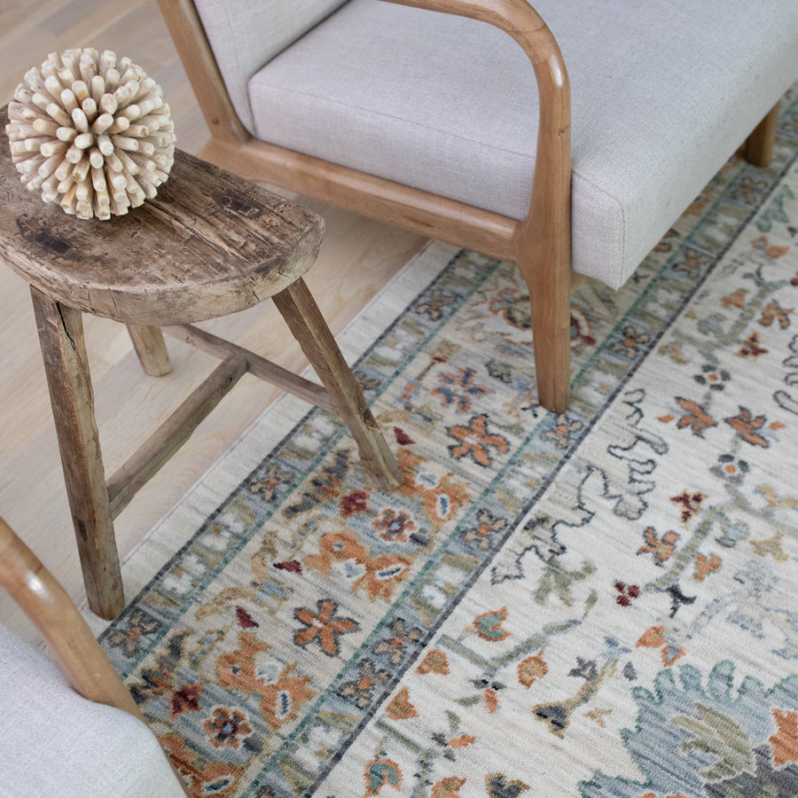 A close-up of a patterned rug with floral motifs in cream, orange, and blue tones, bordered by two light wooden chairs and a rustic wooden stool holding a decorative wooden sphere.