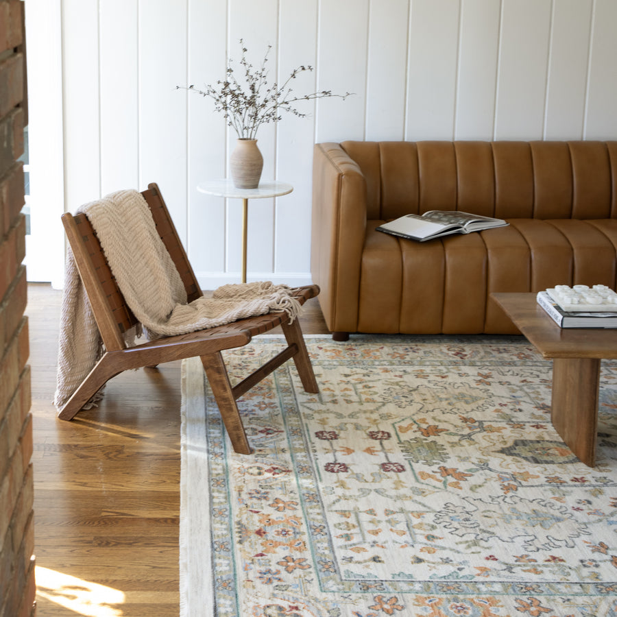 A cozy living room with a tan leather sofa, an open magazine, a wooden chair draped with a beige throw, a patterned rug, a wooden coffee table, and a vase with branches on a small round side table.
