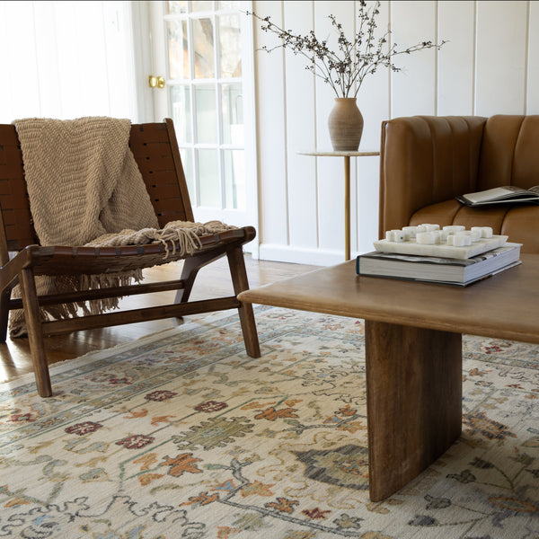 A cozy living room with a wooden chair draped in a knitted blanket, a tan leather sofa, a wooden coffee table, and a side table with a vase of branches. The space features a patterned rug and white-paneled walls.