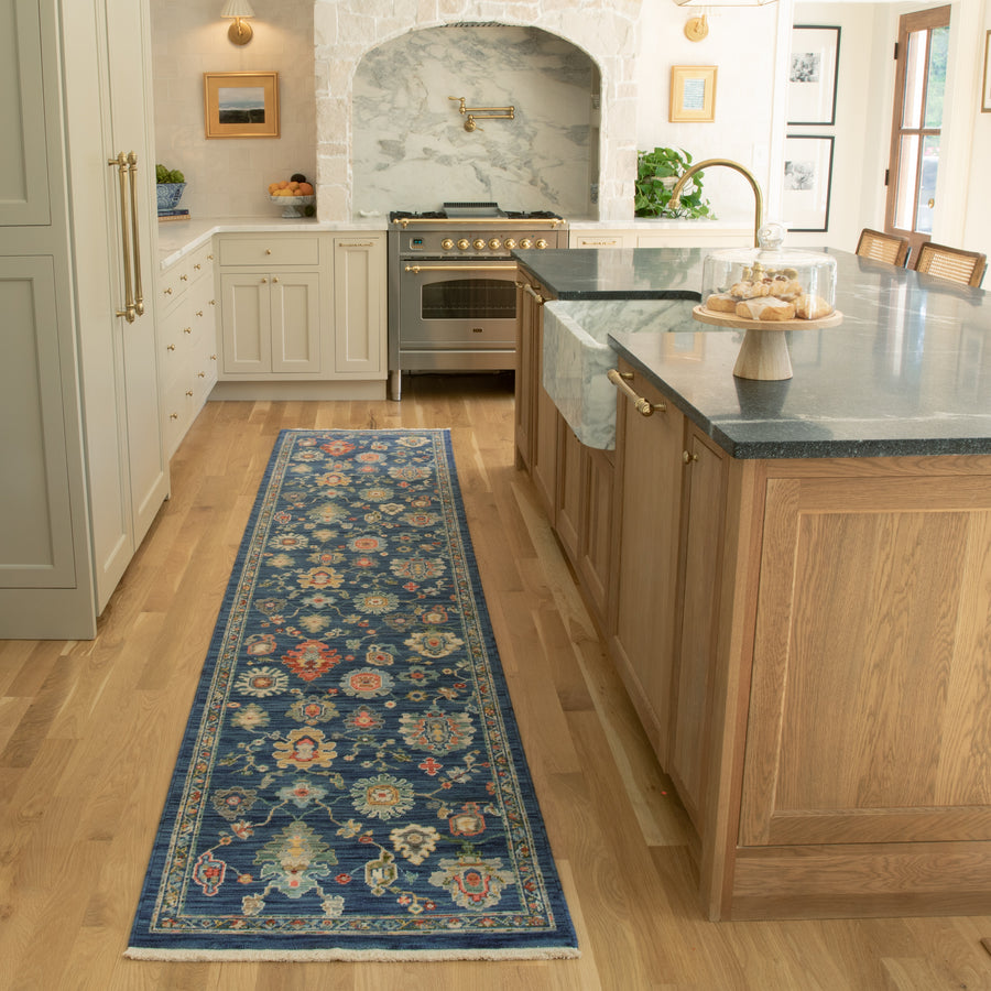 A bright kitchen with light wood floors, white cabinets, a marble island, a stainless steel stove, and a long blue floral runner rug. A cake stand with pastries is on the island.