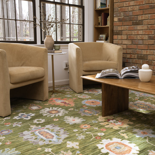 Two tan armchairs sit beside a wooden coffee table with an open book on it, atop a green patterned rug. A vase with branches is on a small side table near large windows and a brick wall.