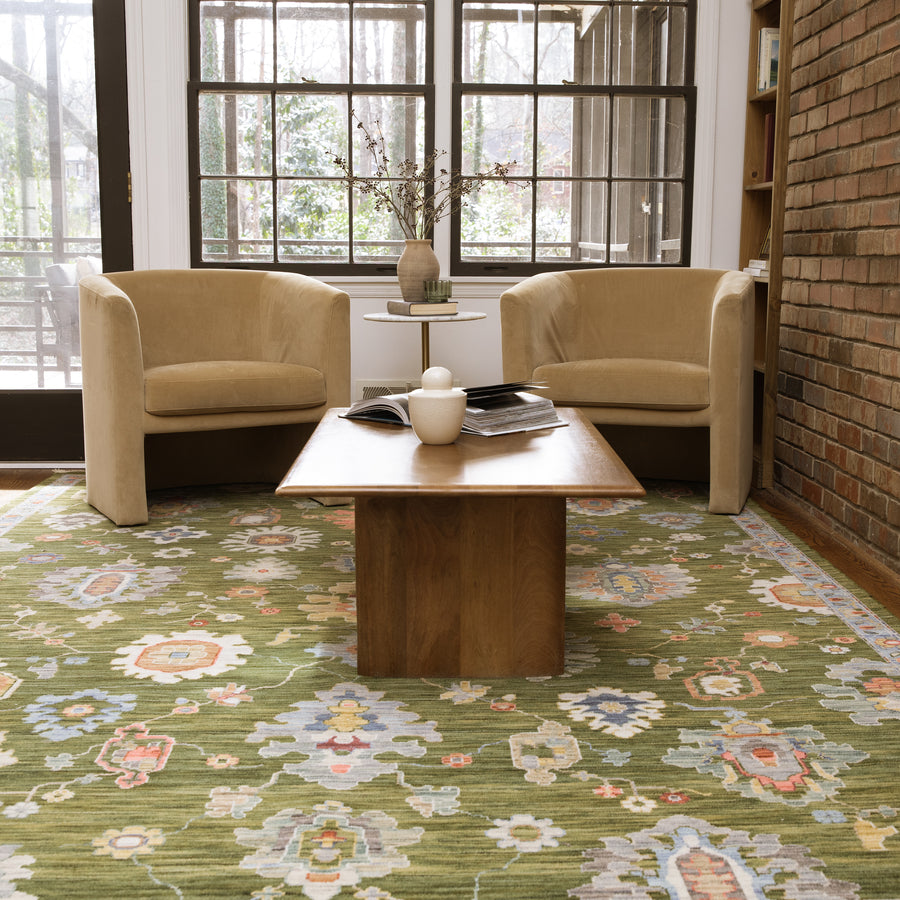 Two tan armchairs face a wooden coffee table with books and a ceramic object, set on a green floral rug in a bright room with large windows and an exposed brick wall.