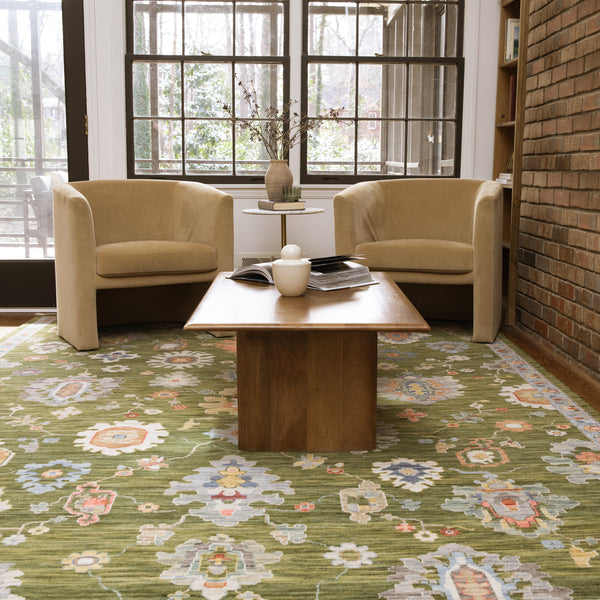 Two tan armchairs face a wooden coffee table with books and a ceramic object, set on a green floral rug in a bright room with large windows and an exposed brick wall.
