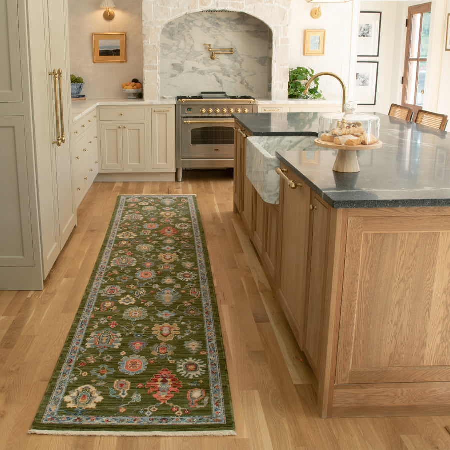 A bright kitchen with light wood floors, a green floral runner rug, beige cabinets, a marble-topped island, and a stainless steel oven set into a stone alcove. A cake stand with pastries sits on the island.