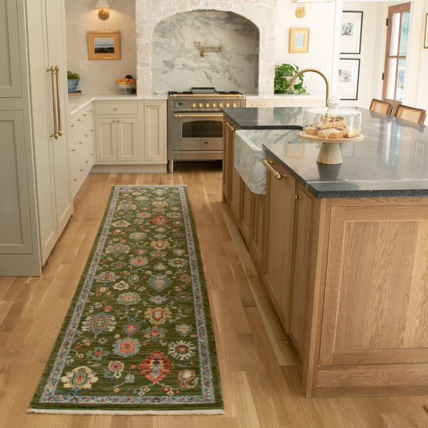 A bright kitchen with light wood floors, a green floral runner rug, beige cabinets, a marble-topped island, and a stainless steel oven set into a stone alcove. A cake stand with pastries sits on the island.