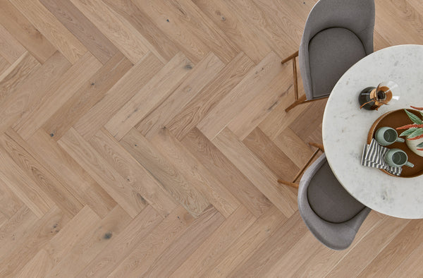 A round white marble table with tableware and plants is next to two gray upholstered chairs on a light wood herringbone-patterned floor, viewed from above.