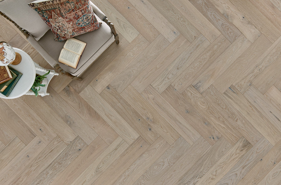 A top view of a beige herringbone wood floor, with a cushioned chair holding a book and patterned pillow, and a small round side table with a teacup and eyeglasses.