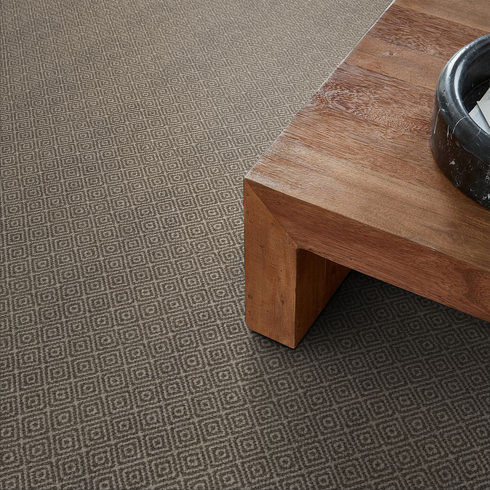 A close-up of a patterned gray carpet with a diamond geometric design, next to the corner of a wooden table and a dark round bowl placed on top of the table.