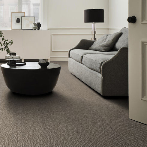A modern living room with a gray sofa, dark round coffee table, white sideboard, black table lamp, and neutral decor, featuring a large window and patterned carpet.