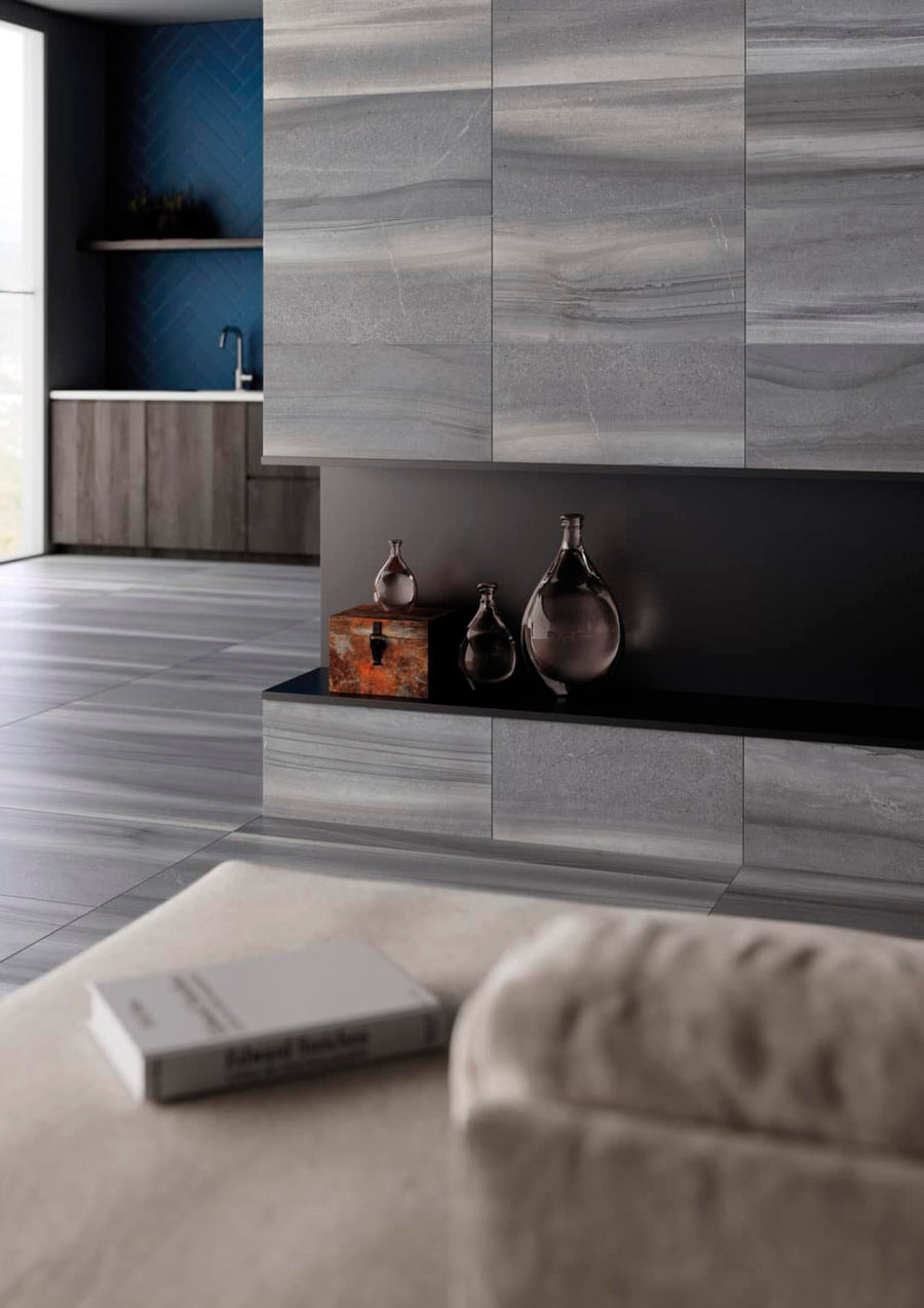 Modern living space with gray stone walls and floors, sleek built-in shelving with decorative glass vases, and a small wooden box. In the background, a kitchen area features dark cabinetry and a deep blue accent wall.