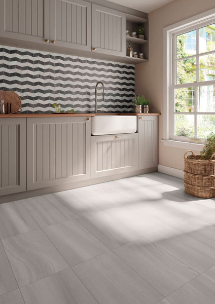 A bright kitchen with gray cabinets, a chevron-patterned backsplash, a farmhouse sink, wooden countertop, and large window letting in sunlight. A woven basket with towels sits on the tiled floor.
