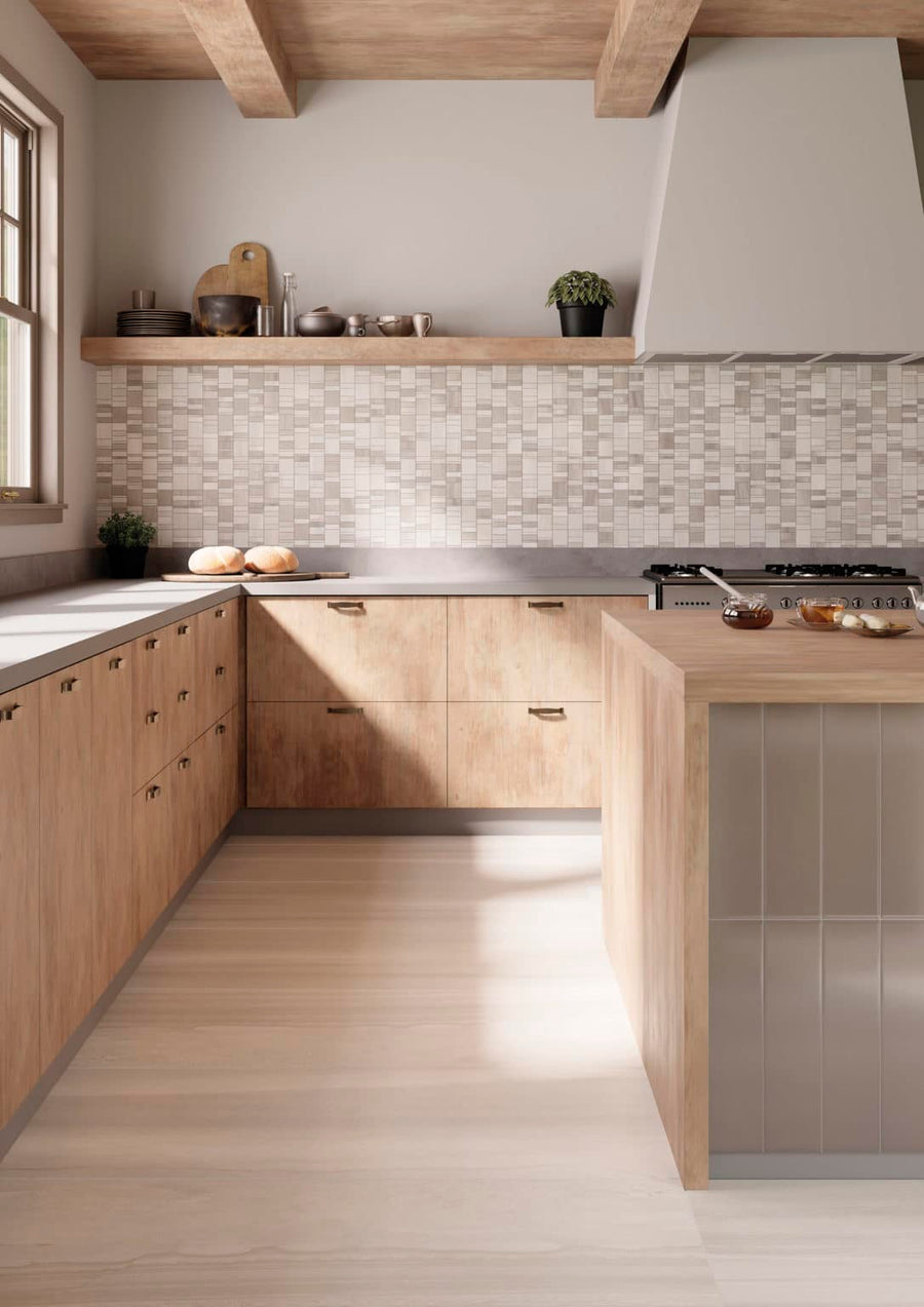 Modern kitchen with light wood cabinets, a gray countertop, a matching island, and a tiled backsplash. There’s a floating shelf with dishes and decor, and sunlight streams in through a window on the left.
