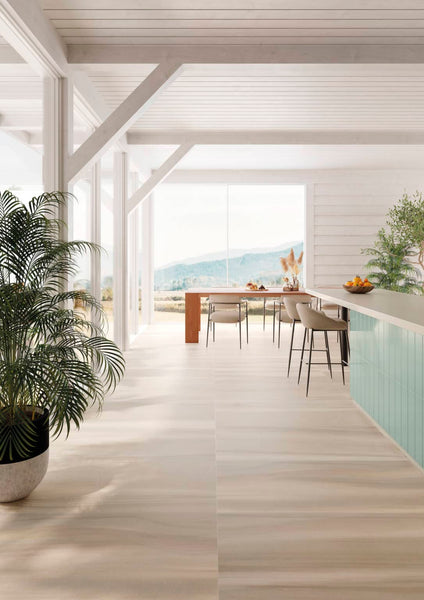 A bright, modern dining area with large windows, light wood flooring, a green kitchen island, potted plants, and a long dining table with chairs, overlooking a scenic outdoor landscape.