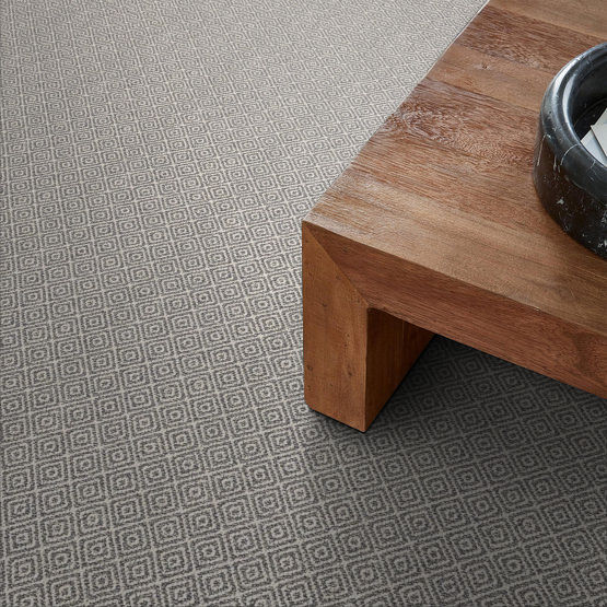 A close-up view of a gray, geometric-patterned carpet next to the corner of a wooden coffee table with a dark round object on top.