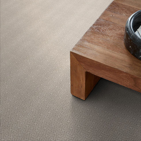 A close-up of a light taupe carpet with a subtle diamond pattern next to a wooden table featuring clean lines. A small dark-colored bowl sits on the table’s corner.
