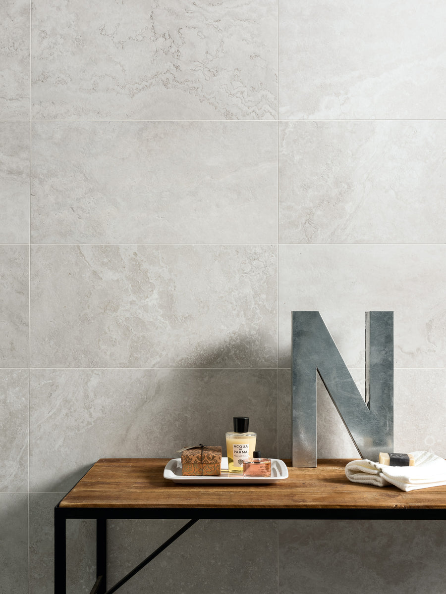 A wooden table against a light stone tile wall holds a metal letter N, a small tray with soap and a lotion bottle, and a folded white towel.