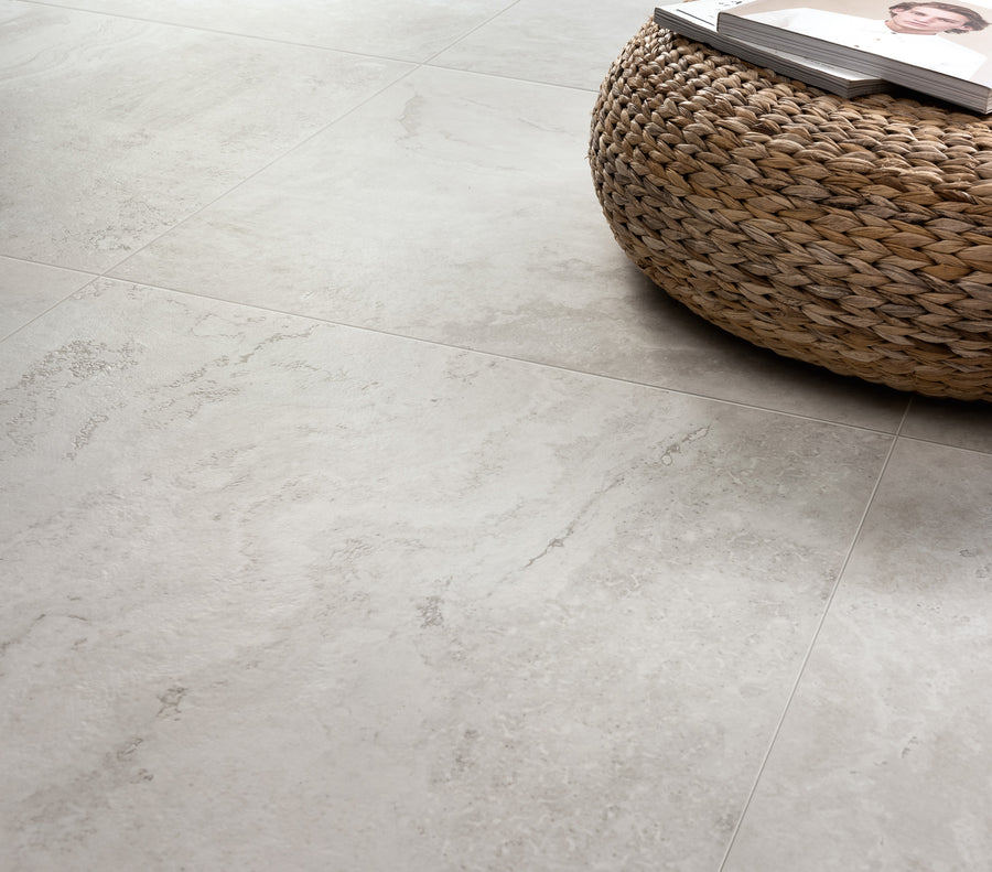 A close-up of light gray, stone-textured floor tiles next to a round, woven wicker table with magazines on top.