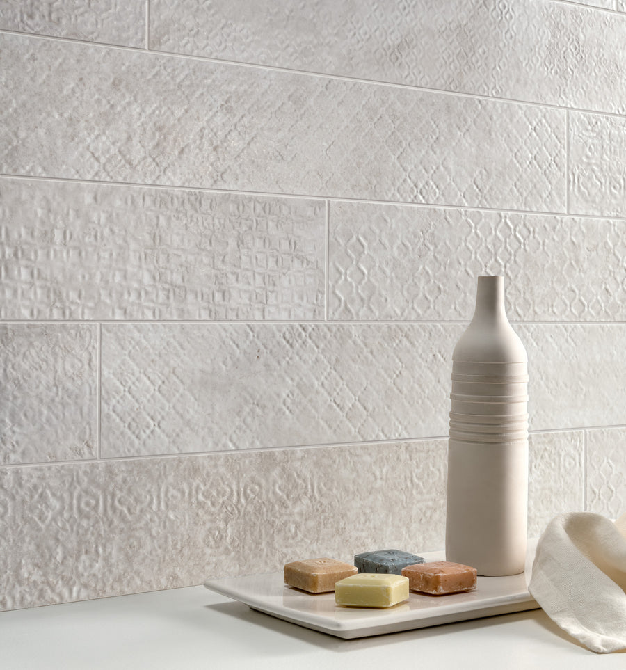 A textured white tile wall, a modern off-white ceramic bottle, and a rectangular plate holding four small, colorful soap bars on a smooth white surface. A cream-colored cloth lies nearby.