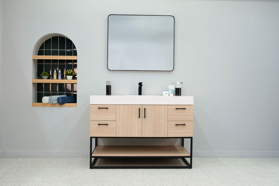A modern bathroom features the Solana 48" Vanity with a black faucet and square mirror above. On the left, a recessed arched shelf displays toiletries, towels, and plants set against dark tile.