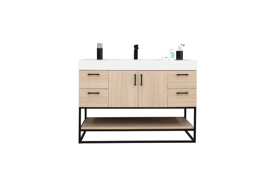 A modern bathroom vanity with a white countertop, black faucet, light wood drawers and cabinets, black handles, and an open shelf underneath, with toiletries placed on top.