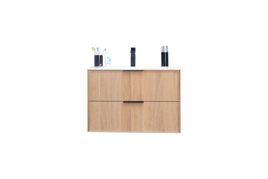 A light wood bathroom vanity with two drawers, a white countertop, and a black faucet. On the counter are toothbrushes in a cup, soap, and two black bottles. The image background is plain white.