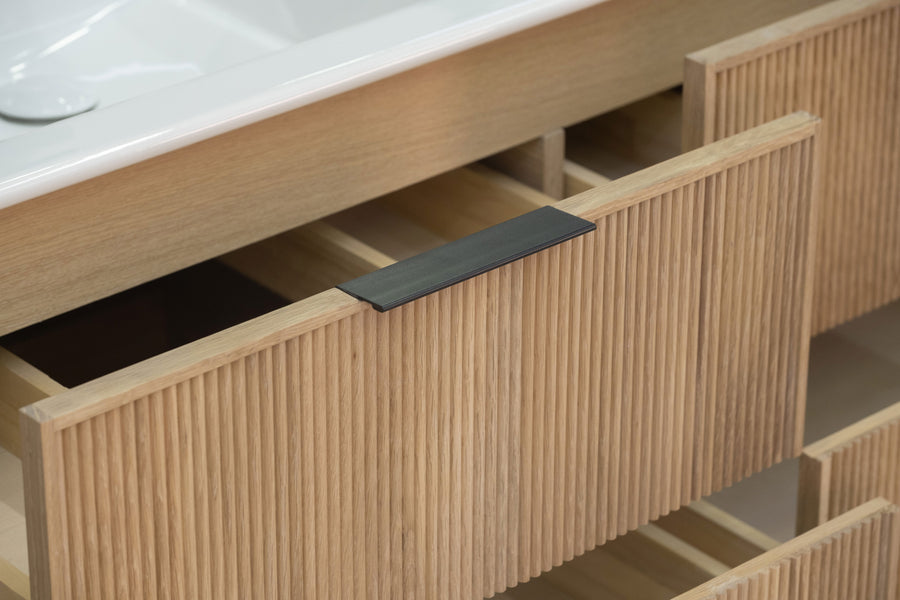 A close-up of a wooden bathroom vanity with open drawers, featuring vertical grooves and a black rectangular handle on the drawer front. The countertop and part of a white sink are visible above.
