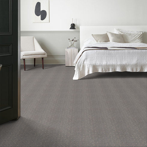 A modern bedroom with a white bedspread, light wood chair, small table with flowers, minimalist wall art, and a grey patterned carpet. The room features neutral tones and simple, elegant decor.