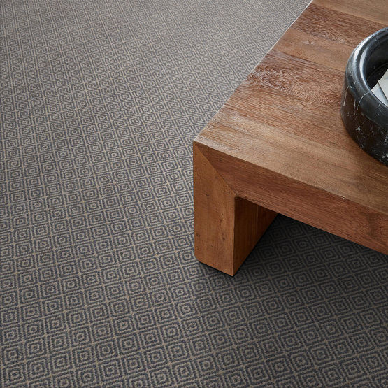 A close-up of a wooden coffee table on a carpet with a small geometric diamond pattern in grey tones. A round, dark-colored dish or bowl sits on the tables corner.