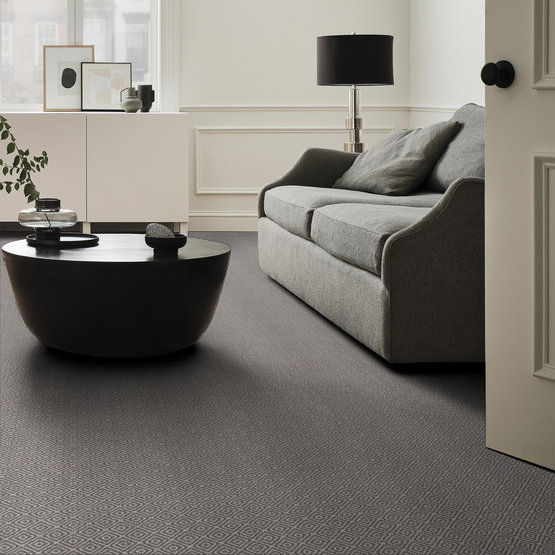 A modern living room with a grey sofa, a round black coffee table, a white sideboard with decor, a black lamp, and wall art. The room has neutral tones and carpeted flooring, with natural light from a window.