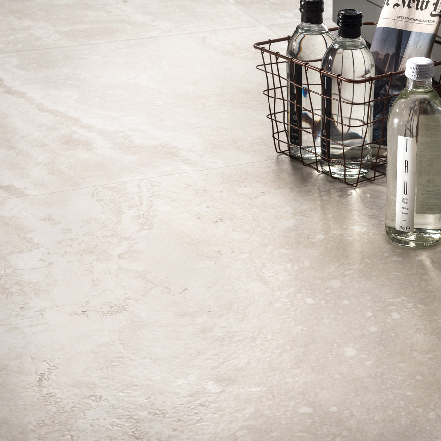 A wire basket holds clear glass bottles and a rolled newspaper on a light beige textured tile floor with a subtle, natural stone pattern.