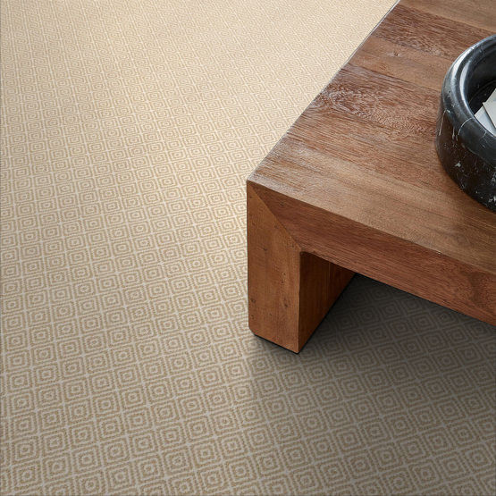 A close-up view of a beige carpet with a subtle diamond pattern next to the corner of a wooden coffee table with a black bowl on top.