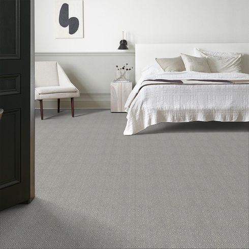 A modern, minimalist bedroom with a neatly made bed, white bedding, a light-colored cushioned chair, small bedside table with a vase, wall art, and a lamp. The room features light gray carpeting and neutral walls.