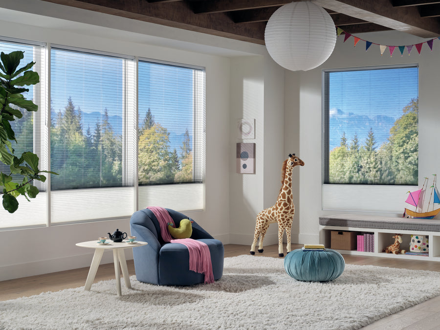 Bright, modern children’s playroom with large windows featuring Applause® Honeycomb Shades, a plush white rug, blue chair with pink blanket, giraffe toy, teal ottoman, and colorful bunting. Trees and mountains are visible outside.