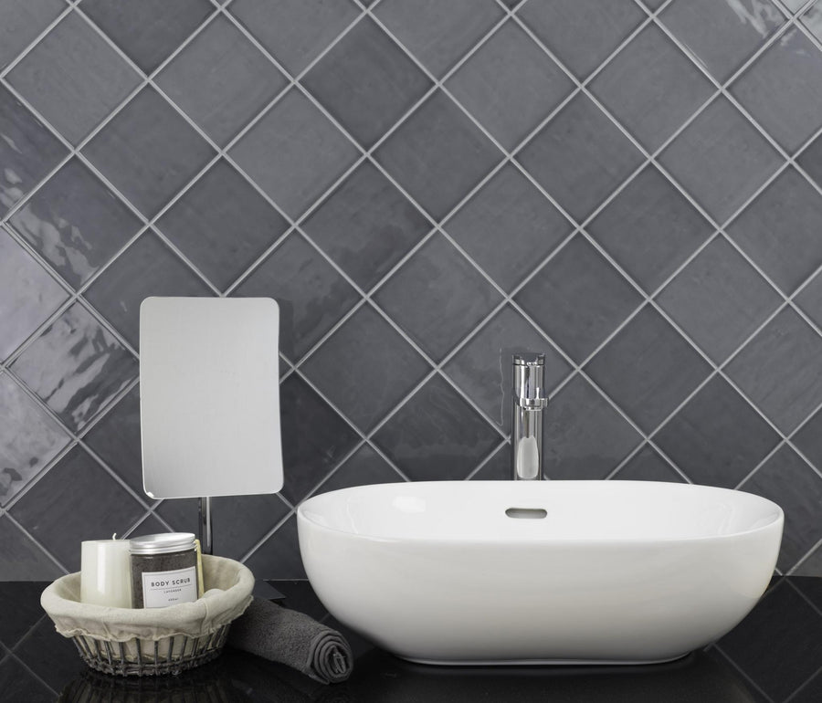 A modern bathroom sink with a silver faucet, small rectangular mirror, grey rolled towel, and a basket holding a candle and body scrub, set against a wall featuring glossy Habitat 5.1x5.1 Subway Tile.