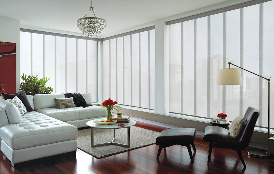 Modern living room with large floor-to-ceiling windows featuring Skyline® Gliding Window Panels, a white sectional sofa, round coffee table with red flowers, black lounge chair and ottoman, floor lamp, hardwood floors, and cityscape views beyond.