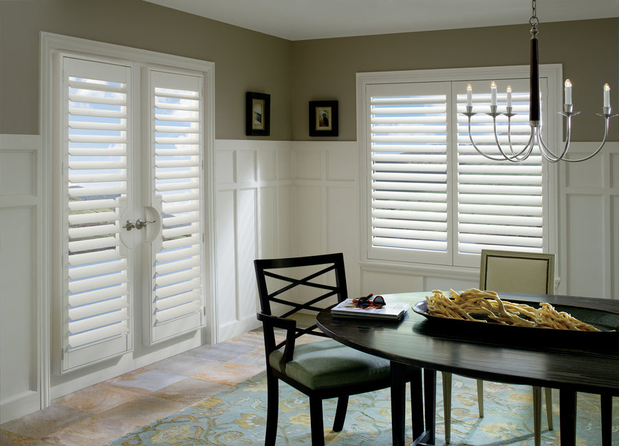 A dining room with elegant UV-resistant Palm Beach™ Polysatin™ Shutters on a window and double doors, a dark wood table with chairs, a modern chandelier, a decorative bowl, and two small framed pictures on the wall.