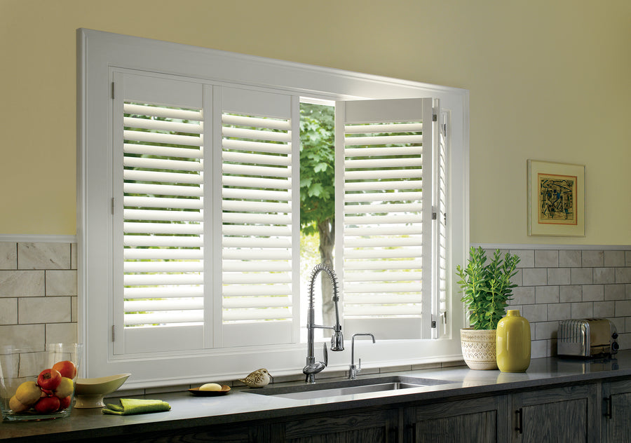 A kitchen window features white Palm Beach™ Polysatin™ Shutters, partially open to show a green tree outside. Below, the countertop includes a sink, faucet, plant, fruit, yellow jar, and a framed picture on the wall.