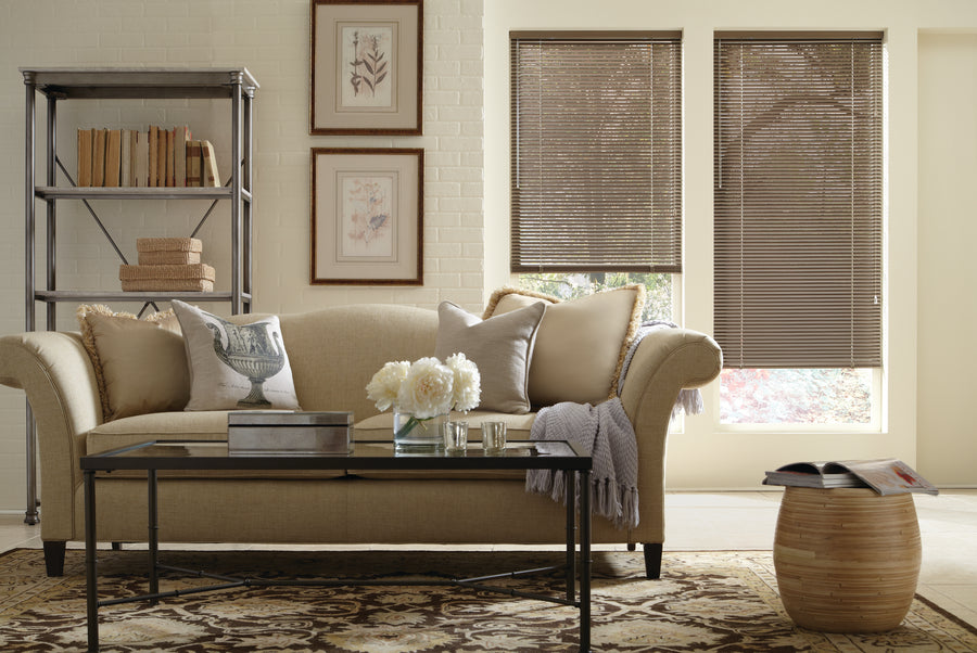 A cozy living room features a beige sofa, neutral pillows, a glass coffee table with books and flowers, a patterned rug, wall art, shelves with books, and windows dressed in Modern Precious Metals® Aluminum Blinds for stylish light control.