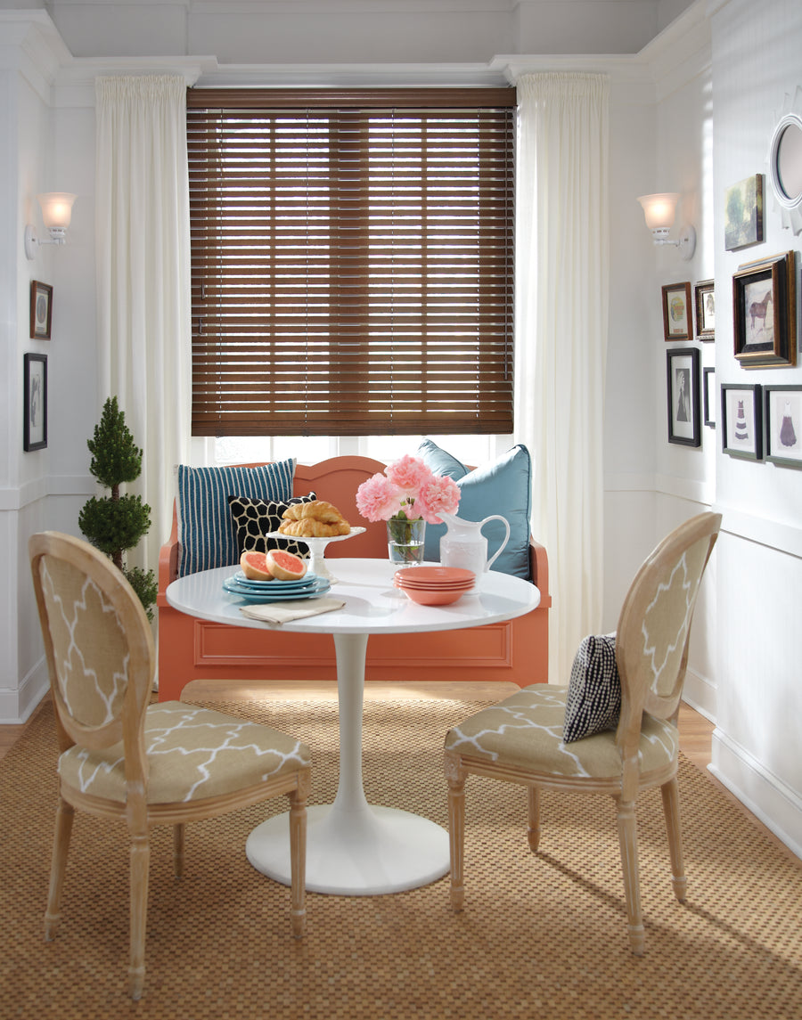 A cozy dining nook features two beige patterned chairs, a white round table with watermelon, croissants, and pink flowers. An orange bench with blue pillows sits beneath a window adorned with Parkland® Wood Blinds and white curtains.